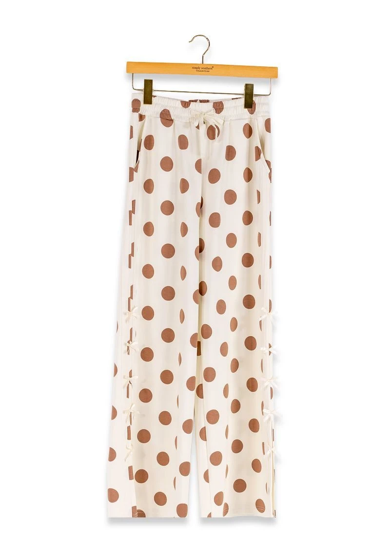 A pair of wide-leg trousers with a brown polka dot pattern on a cream background, hanging from a wooden hanger against a white background.