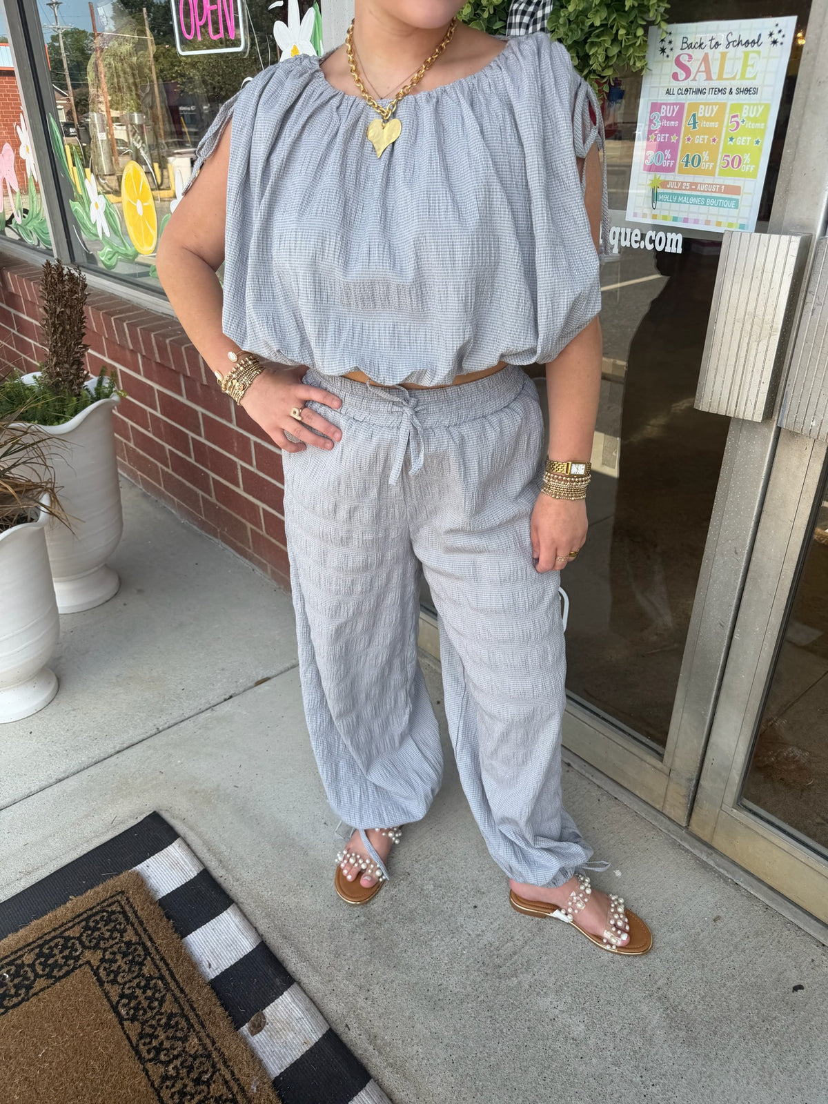 A person wearing a light blue-gray crinkle fabric two-piece set with a cropped off-the-shoulder top and matching wide-leg pants, standing on a sidewalk in front of a storefront.