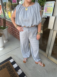 A person wearing a light blue-gray crinkle fabric two-piece set with a cropped off-the-shoulder top and matching wide-leg pants, standing on a sidewalk in front of a storefront.