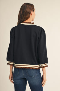 A woman shown from the back wearing a black cropped jacket with brown and cream striped ruffle trim at the collar, cuffs, and hem, paired with blue jeans against a light beige studio background.