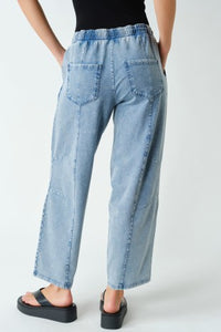 A person shown from the back wearing light-wash blue wide-leg denim jeans with an elastic waistband, patch pockets, and a raw hem, paired with black platform sandals against a plain light background.