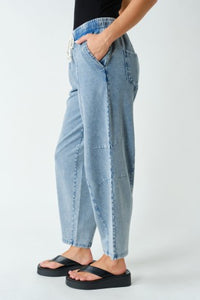 A person wearing light-wash blue denim pants with a relaxed, baggy fit and black flip-flop sandals, shown from the waist down against a plain light gray studio background.