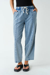A person wearing light-wash blue cropped denim pants with a white drawstring waist, standing against a plain light gray studio background. The person is cropped from the chest down and wearing black sandals.