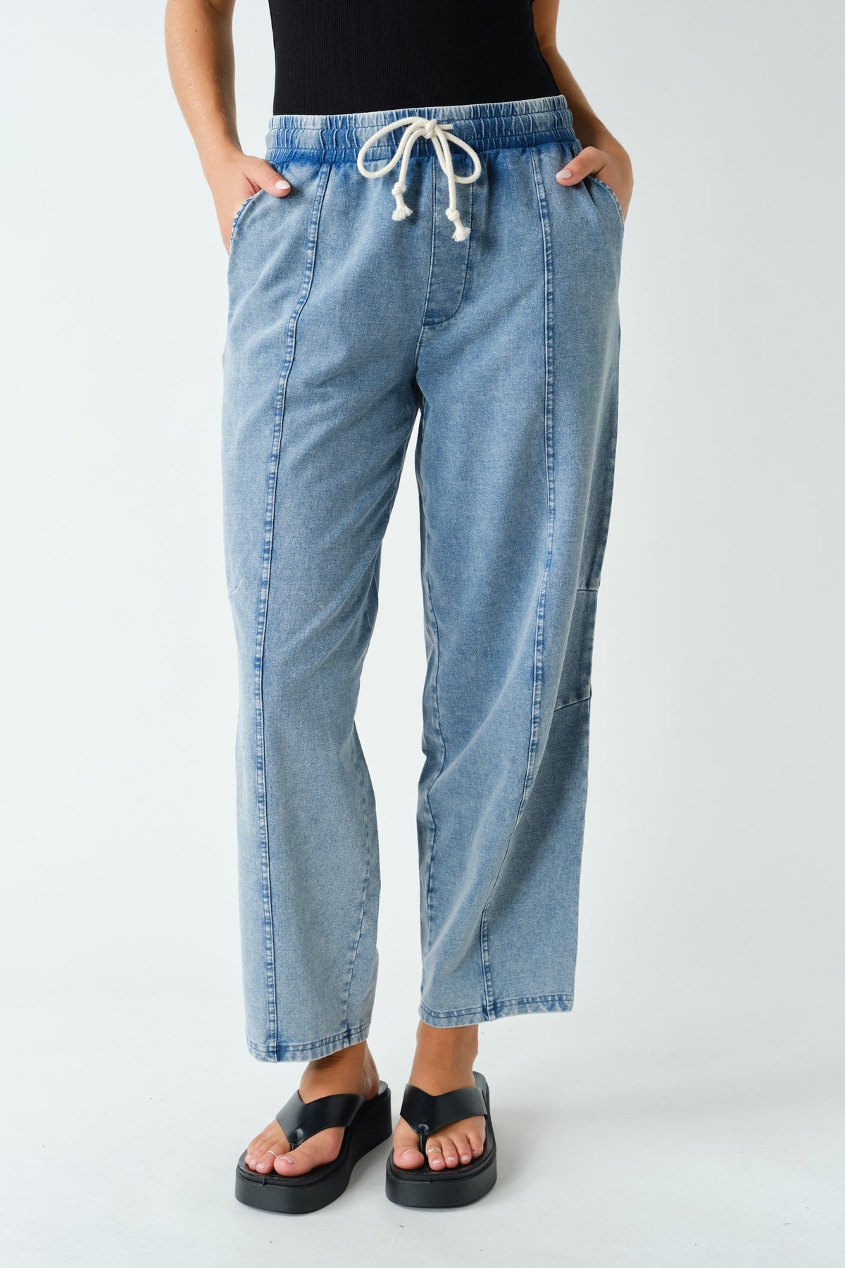 A person wearing light-wash blue cropped denim pants with a white drawstring waist, standing against a plain light gray studio background. The person is cropped from the chest down and wearing black sandals.