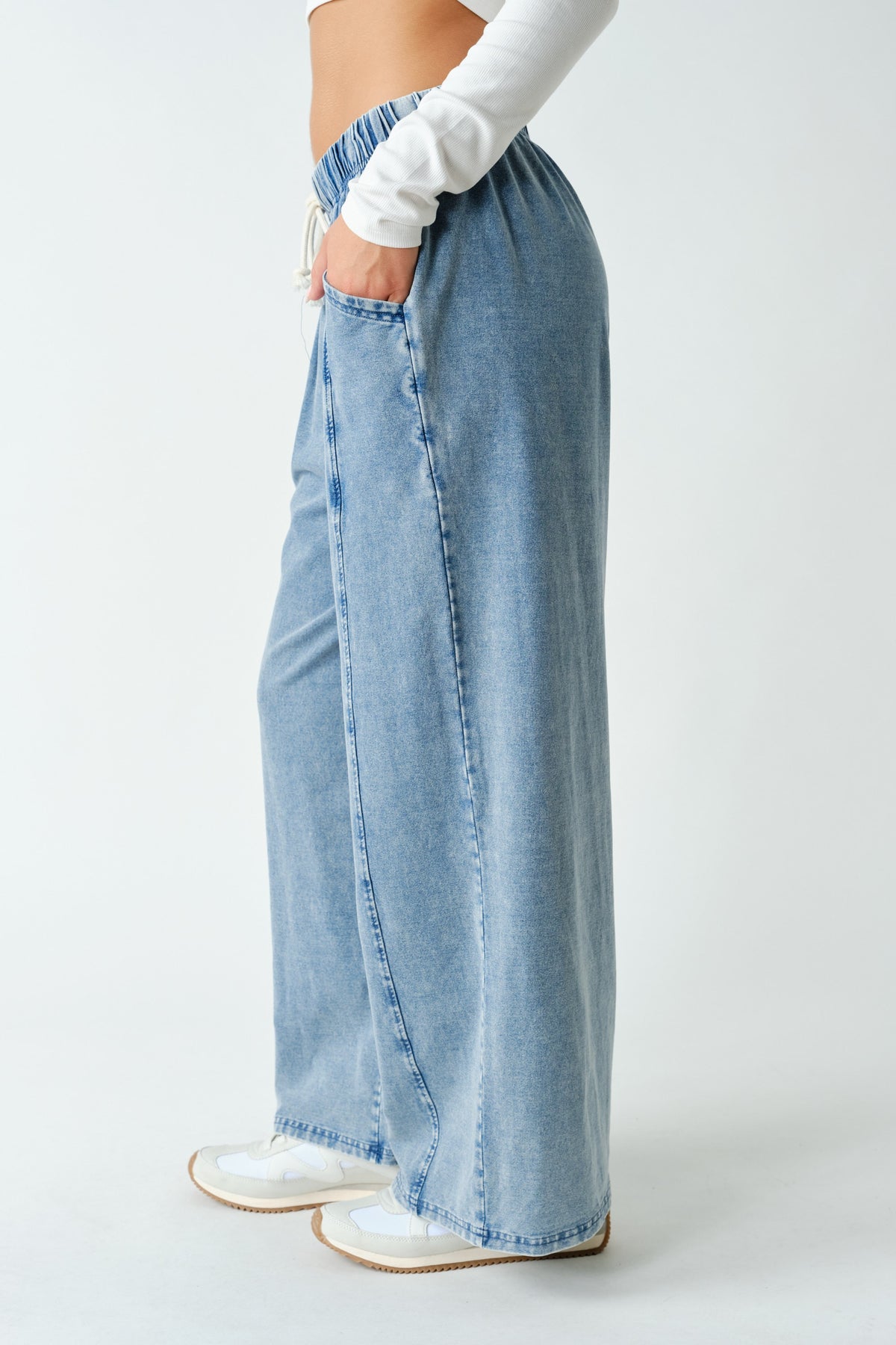 A person wearing light-wash wide-leg denim pants with side pockets, paired with a white long-sleeve top and white sneakers, shown from the waist down against a plain light gray studio background.