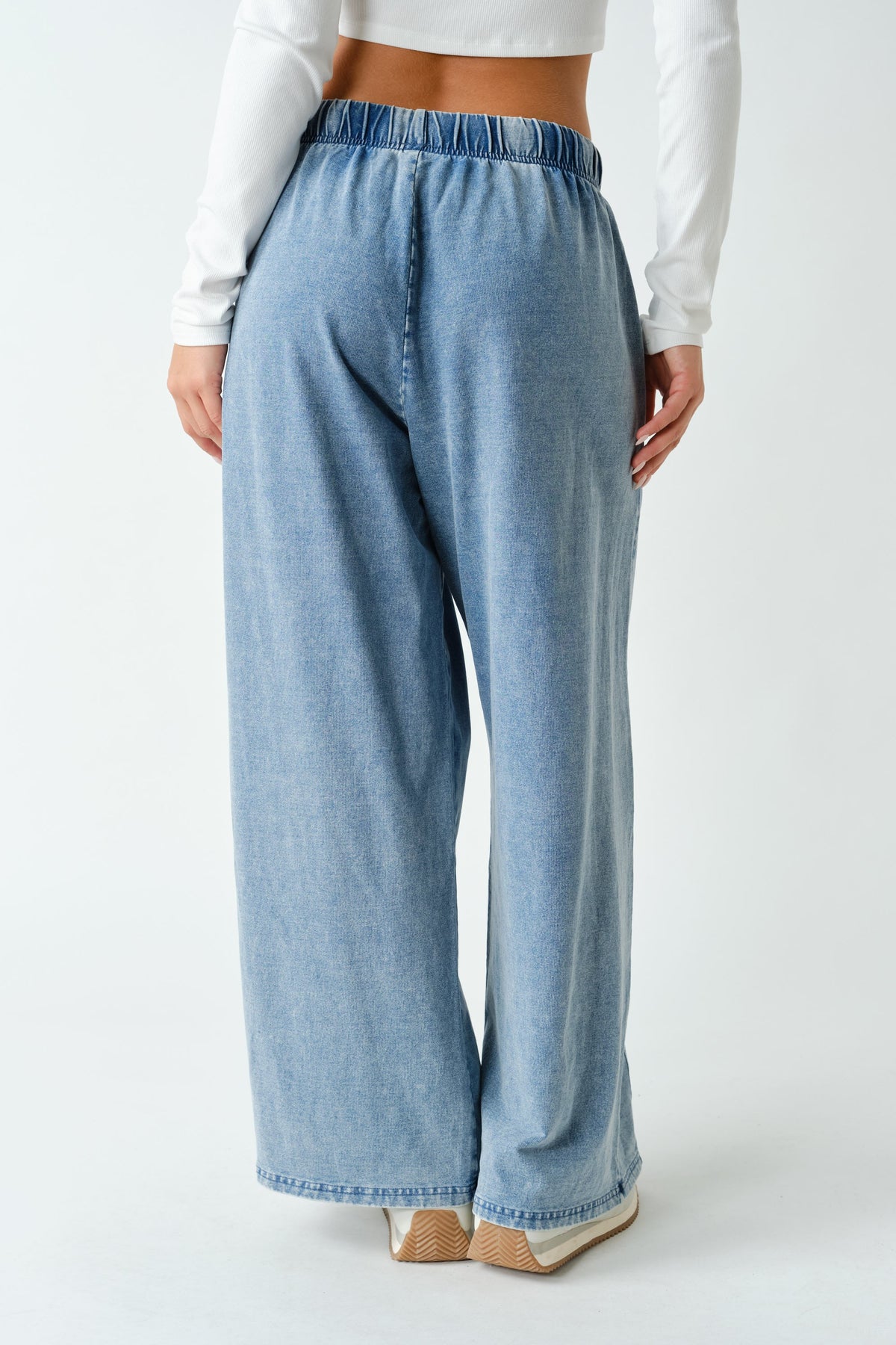 A person shown from the back wearing light blue wide-leg denim pants with an elastic waistband, paired with a white long-sleeve crop top and beige heels, against a plain light gray studio background.