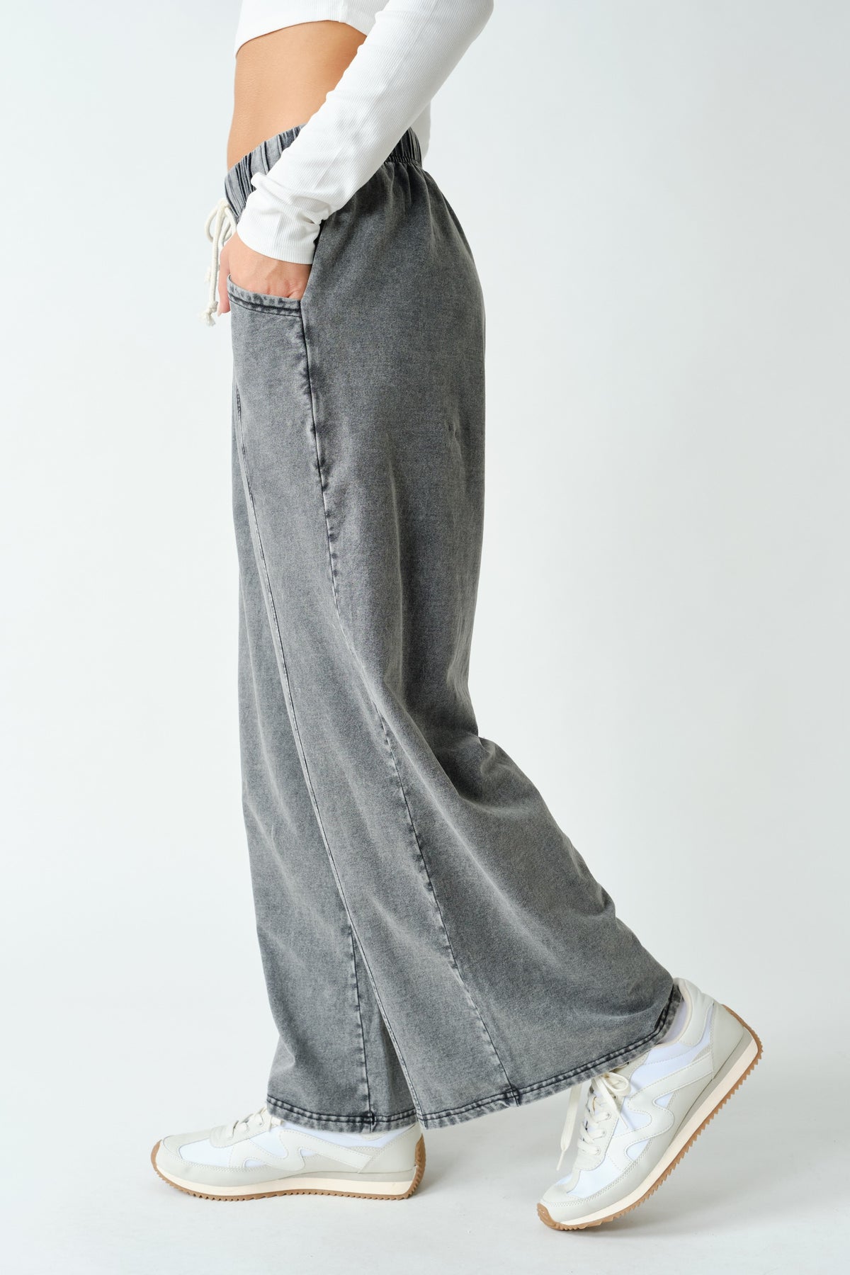 A person wearing wide-leg gray denim jeans with a faded wash, styled with a white long-sleeve crop top and white sneakers, shown from the waist down against a light gray studio background.