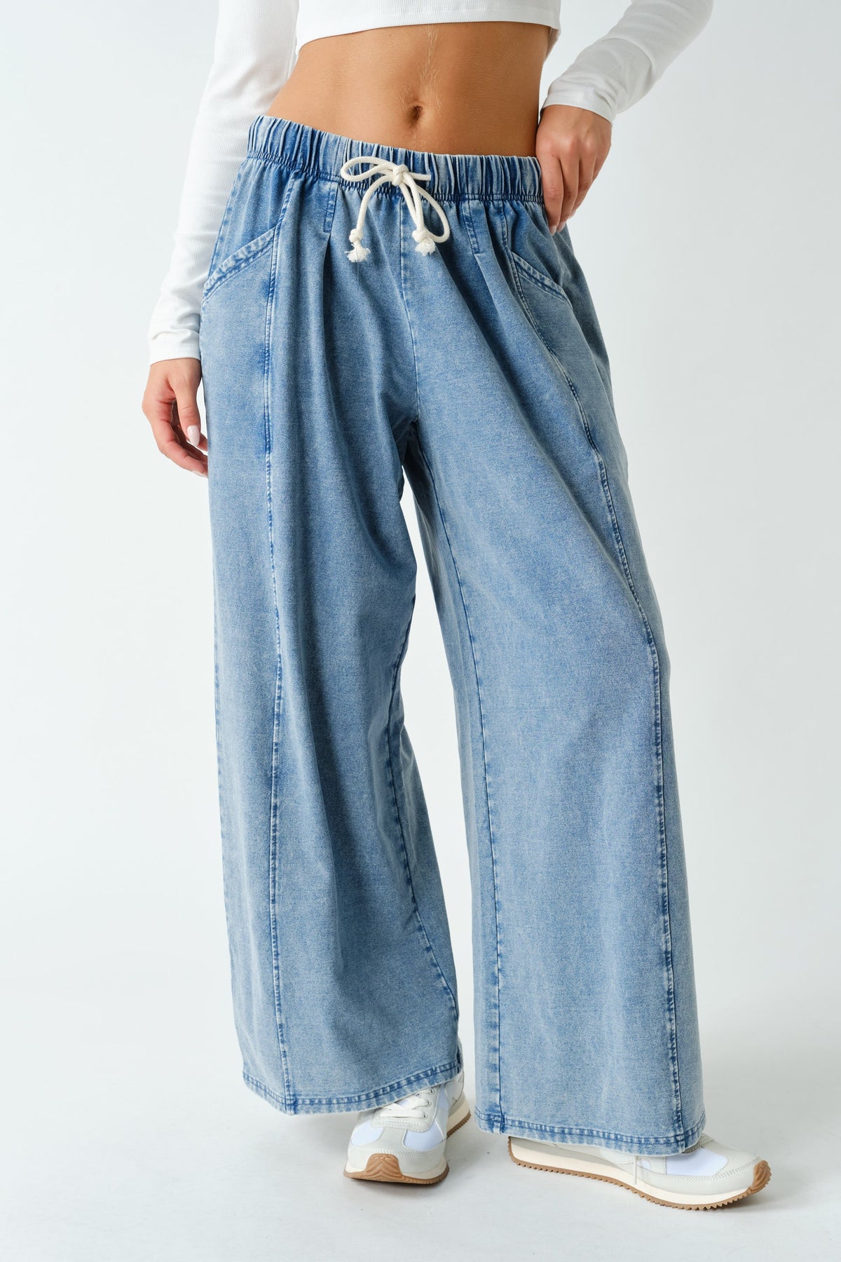 A person wearing light blue wide-leg denim jeans with an elastic waistband and white drawstring, paired with a white long-sleeve top and white sneakers, against a plain light gray studio background.