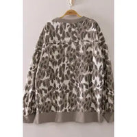 A long-sleeve crewneck sweatshirt with an oversized leopard print pattern in gray, brown, and beige, hanging on a wooden hanger against a plain light gray wall.