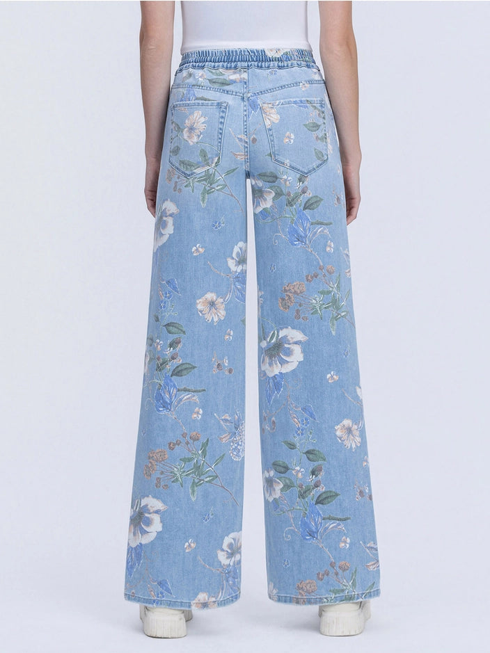 A person shown from the back wearing light blue wide-leg denim jeans with an allover floral print in white, blue, and green, paired with a white top and white sneakers against a light gray studio background.