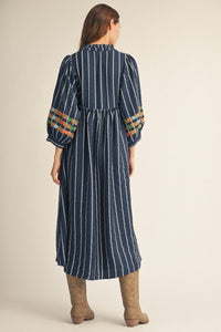 A woman viewed from the back wearing a navy blue midi dress with white vertical stripes, gathered elbow-length puff sleeves, and colorful embroidered details near the cuffs. She is wearing tan boots against a plain beige studio background.