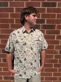 A man wearing a short-sleeve button-up shirt with a beige, brown, and gray camouflage pattern, standing against a red brick wall.