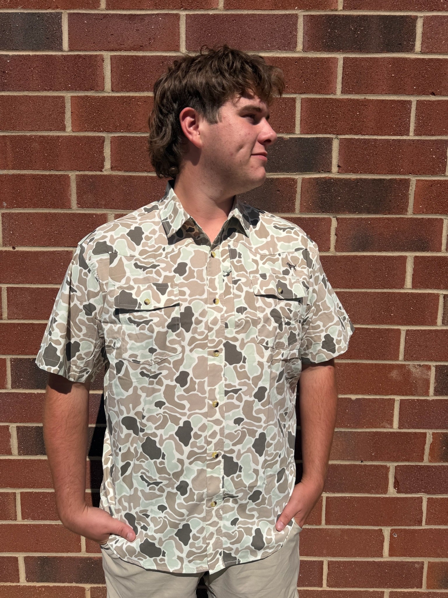 A man wearing a short-sleeve button-up shirt with a beige, brown, and gray camouflage pattern, standing against a red brick wall.