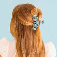 A woman with long light brown hair is shown from the back, wearing a patterned hair claw clip that holds a half-up bun. The background is a solid light blue.