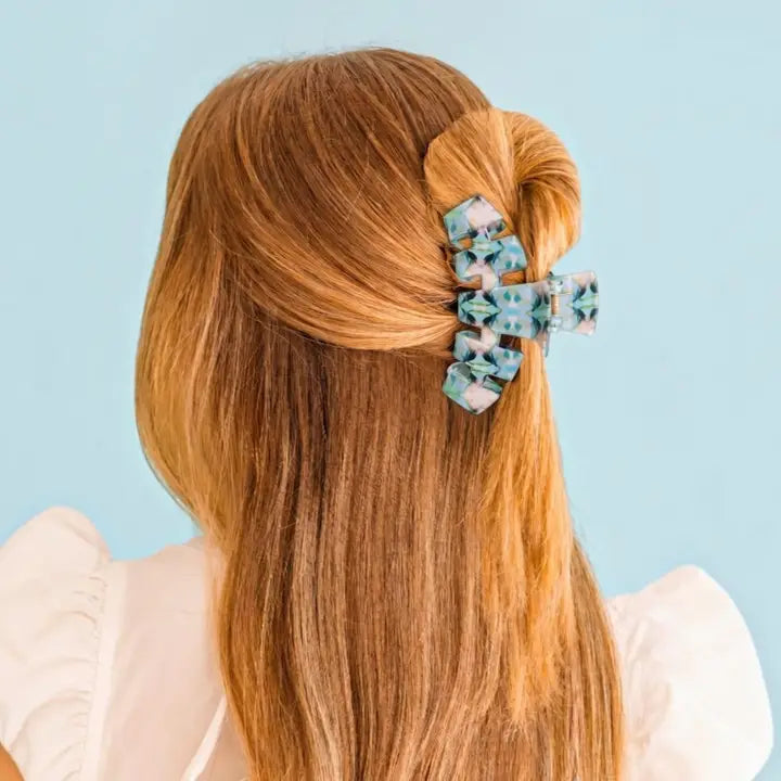 A woman with long light brown hair is shown from the back, wearing a patterned hair claw clip that holds a half-up bun. The background is a solid light blue.