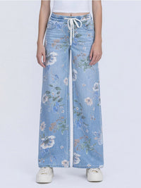 A model is wearing a pair of wide-leg light blue denim jeans with a white floral print and a white drawstring waist, paired with white sneakers against a light gray background.