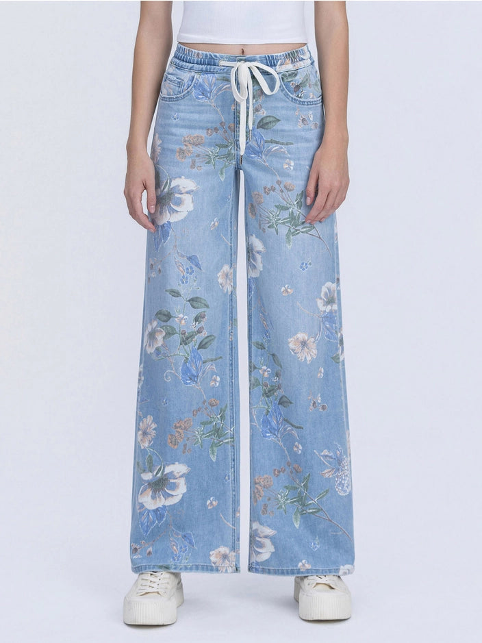 A model is wearing a pair of wide-leg light blue denim jeans with a white floral print and a white drawstring waist, paired with white sneakers against a light gray background.