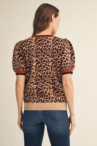A woman viewed from the back wearing a leopard-print short-sleeve top with puff sleeves and contrasting ribbed trim, paired with blue jeans, against a plain beige studio background.