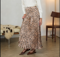 A person wearing a pleated midi skirt with a beige, brown, and black leopard print, styled with a white long-sleeve top and brown pointed flats in a modern indoor setting.