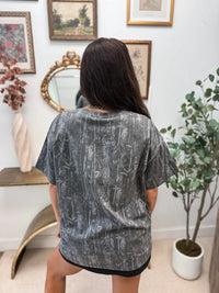 A woman viewed from behind wearing an oversized short-sleeve t-shirt with a gray and black abstract brushstroke pattern, standing in a decorated room with framed art, a mirror, and a potted plant.