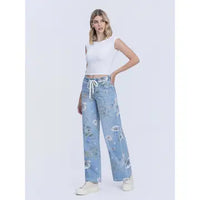 A woman modeling light blue wide-leg jeans with a floral print and white drawstring waist, paired with a white sleeveless crop top and white sneakers, against a light gray studio background.