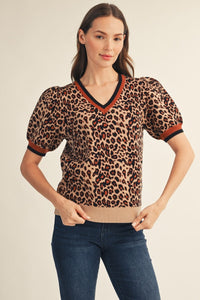 A woman modeling a short-sleeve leopard-print knit top with puff sleeves and a V-neck, paired with dark blue jeans against a plain light beige studio background.