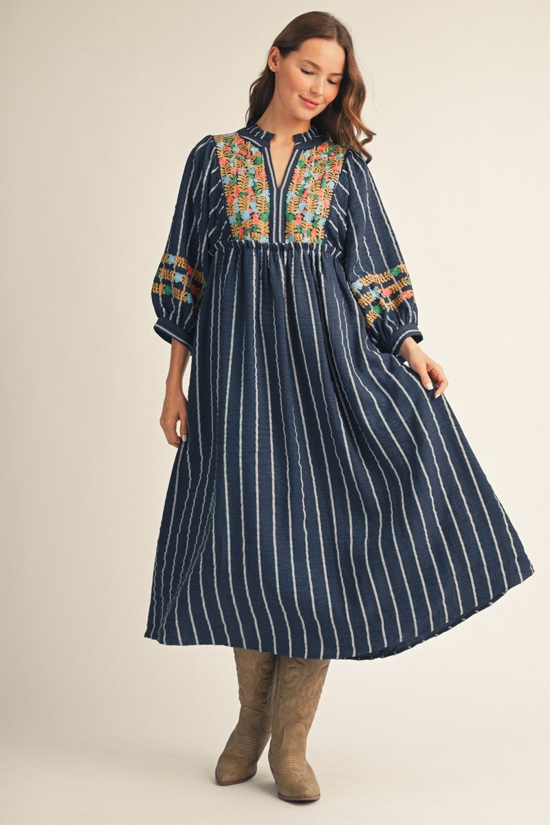 A woman modeling a navy blue maxi dress with vertical white stripes, a colorful embroidered yoke, and patterned sleeve cuffs, paired with brown boots against a light beige studio background.