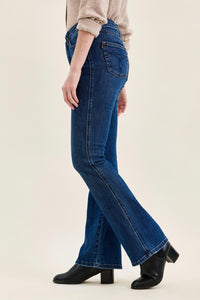 A side-profile view of a person wearing dark blue high-rise flare jeans, a beige long-sleeve top, and black ankle boots against a plain light beige studio background.