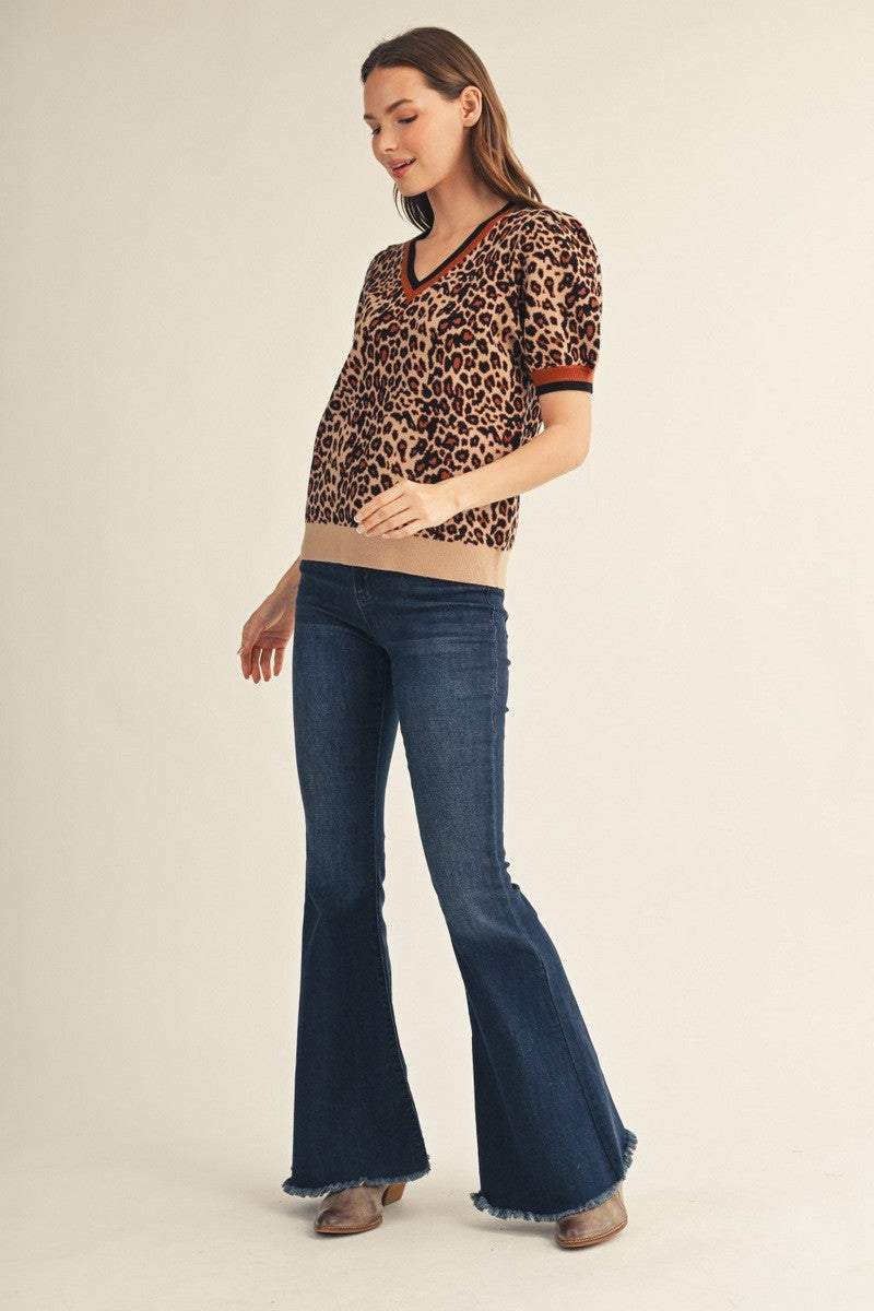 A woman modeling a short-sleeve leopard-print knit top with dark red trim and dark blue flare jeans against a plain beige studio background.