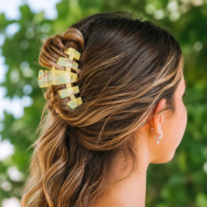 A woman shown from the side with her hair in a half-up bun secured by a large rectangular claw hair clip. The clip has a light yellow-green base with a distressed, mottled pattern in muted tones.
