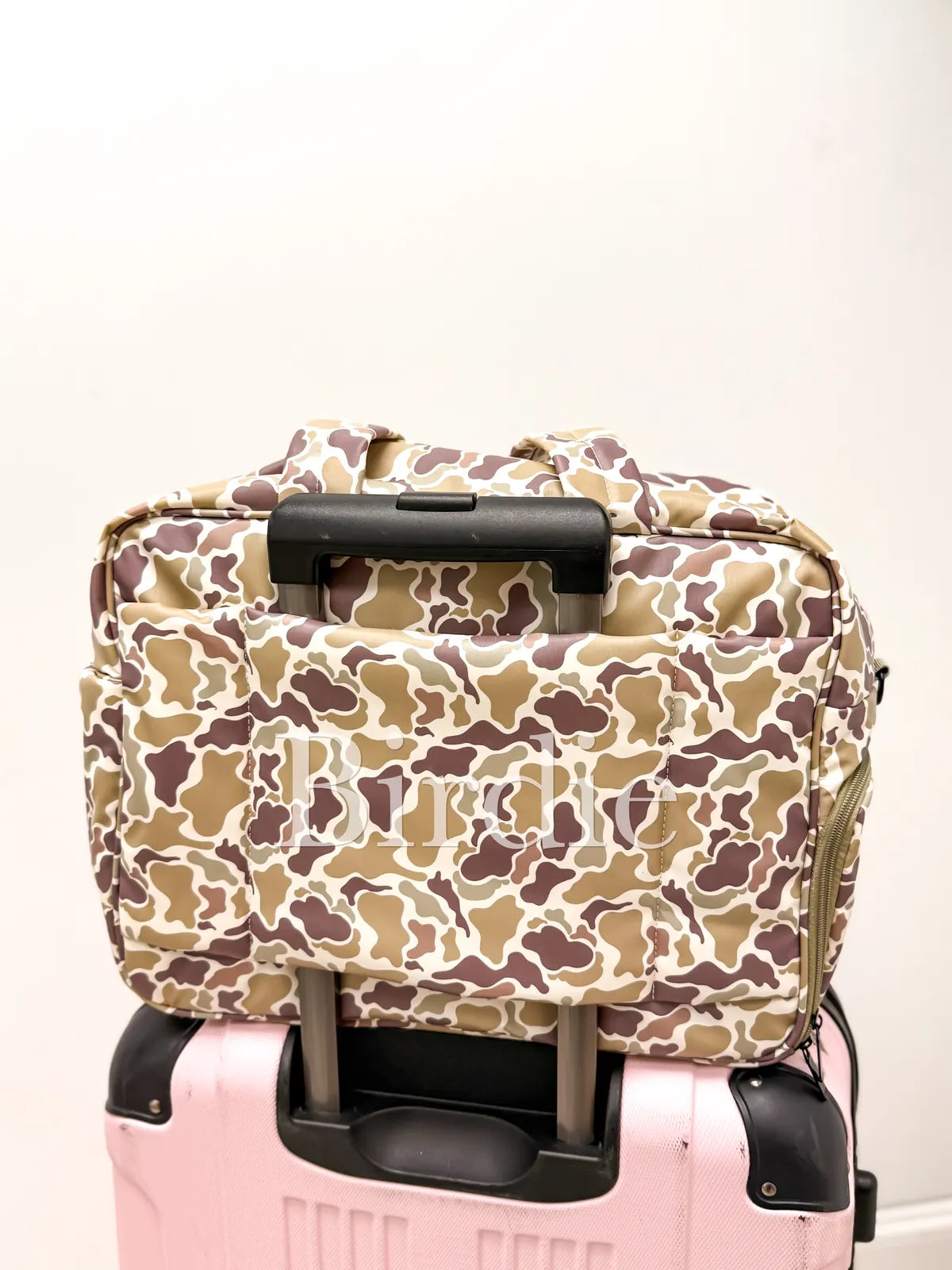 A beige, brown, and white camouflage patterned soft-sided bag with black handles, stacked on top of a pink hard-shell suitcase against a plain off-white wall.