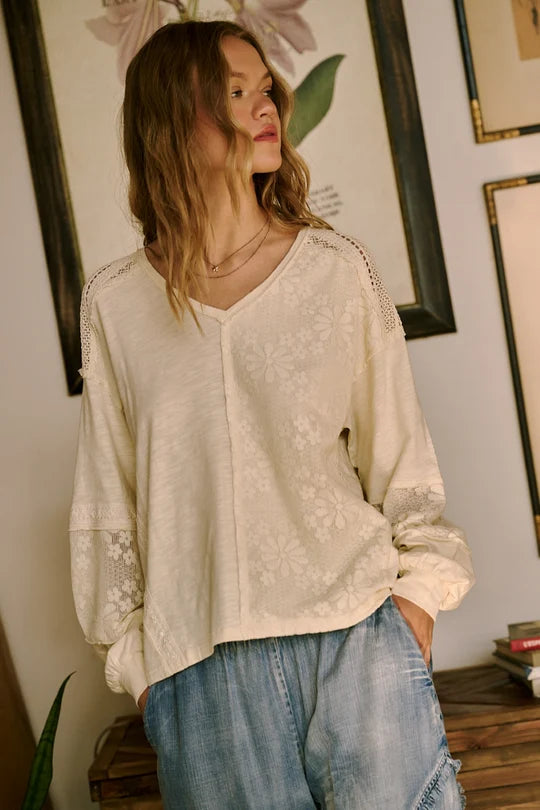 A woman wearing a cream-colored, long-sleeve V-neck blouse with delicate floral embroidery and lace details, paired with light-wash blue jeans, standing in a room with framed wall art.